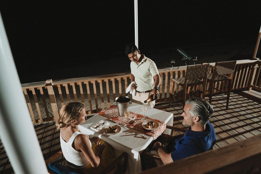 A waiter pours champagne for a man and a woman enjoying dinner on a terrace under the night sky.